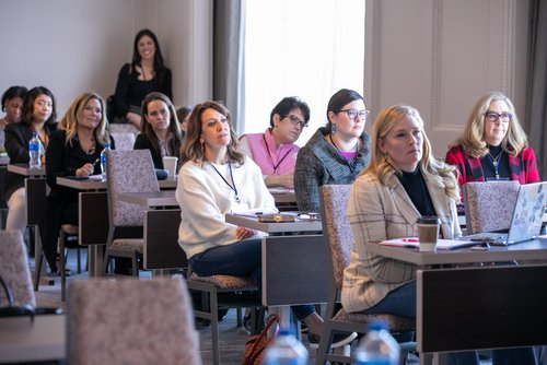 A large group of women sit at desks, focusing intently on the front of the room.