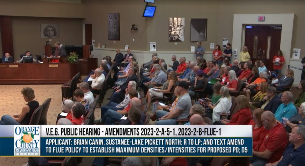 Room full of residents seated at the Board of County commission chambers during the Sustanee PD transmittal hearing in District 5. Text reads V.E.6. Public Hearing-Amendmenrs 2023-2-A-5-1,2023-2-B-FLUE-1. Applicant: Brian Canin, Sustanee-Lake Pickett North: R to LP, and Text Amend to FLUE Policy to Establish Maximum Densities/Intensities for Proposed PD; D5.