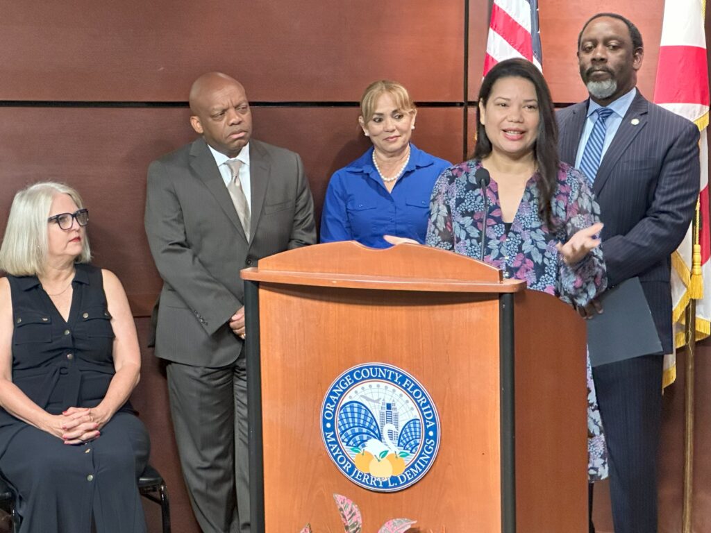 photograph of commissioner bonilla speaking at a lectern, surrounded by the mayor and other commissioners